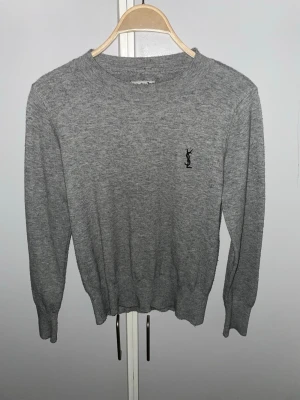 Grå stickad tröja från Yves Saint Laurent  - Snygg grå stickad tröja från Yves Saint Laurent med diskret svart YSL-logga broderad på bröstet. Storlek S, tröjan har rund halsringning, långa ärmar och ribbade muddar vid ärmslut och nederkant. Perfekt för en stilren och enkel look.