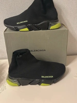 Balenciaga svarta sneakers med neon - Säljer ett par svarta Balenciaga sneakers med stickad ovandel och chunky sula. Skorna har neon-gula detaljer på sulan och Balenciaga-logga i gult på sidan. Modern strumpliknande design som ger en clean och futuristisk vibe.
