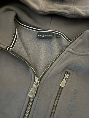 Mörkgrå hoodie från Sail Racing - Säljer en mörkgrå hoodie från Sail Racing med dragkedja framtill och en extra bröstficka med dragkedja. Hoodien har en mjuk insida och broderad logga på bröstet. Perfekt för chill dagar och streetwear-stil.