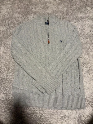 Grå  kabelstickad tröja Polo Ralph Lauren - Ljusgrå kabelstickad tröja från Polo Ralph Lauren med halv dragkedja och hög krage. Tröjan har ribbade muddar och nederkant samt den klassiska blå logotypen broderad på bröstet. Perfekt för lager på lager och en stilren look.