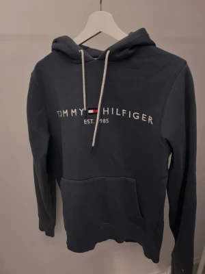 Blå hoodie från Tommy Hilfiger - Mörkblå hoodie från Tommy Hilfiger med vit broderad logga och flagga på bröstet. Klassisk känguruficka framtill, justerbar huva med vita snören och mjukt material i french terry. Perfekt för en avslappnad och stilren look.