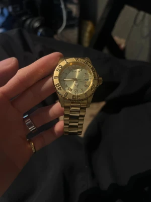 Guldfärgad klocka från Invicta - Snygg guldfärgad klocka från Invicta med metallarmband och klassisk rund urtavla. Klockan har romerska siffror, datumvisning och en robust länkdesign. Perfekt accessoar för dig som gillar en lyxig och stilren look.