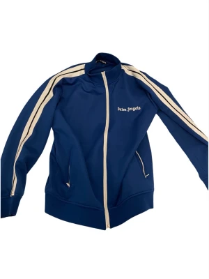 Blå track jacket från Palm Angels - Snygg blå track jacket från Palm Angels med vita ränder längs ärmarna och dragkedja framtill. Jackan har hög krage, två fickor med dragkedja och logga på bröstet. Perfekt för en sportig och trendig look.