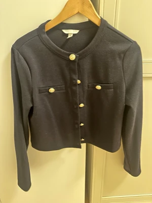 Svart kofta med guldknappar H&M - Stilren svart kofta från H&M med rund halsringning och dekorativa guldknappar framtill. Två fejkfickor med matchande knappar på bröstet. Långärmad och i mjukt, stretchigt material som ger en snygg och klassisk look.