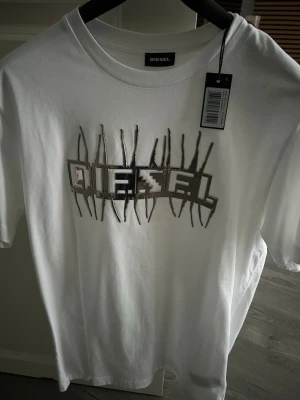 Vit t-shirt med grafiskt tryck från Diesel - Säljer en vit t-shirt från Diesel med ett unikt grafiskt tryck framtill där märkesnamnet är inramat av ett mönster som liknar säkerhetsnålar. Klassisk rund hals och korta ärmar. Perfekt för dig som vill sticka ut med en cool detalj.