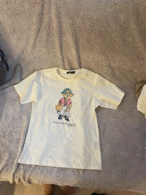 Vit t-shirt Polo PJ by Polo Company - Vit t-shirt med färgglatt tryck av en björn på framsidan från Polo PJ by Polo Company. Klassisk passform med rund hals och korta ärmar. Perfekt för en avslappnad och trendig stil. Älskar