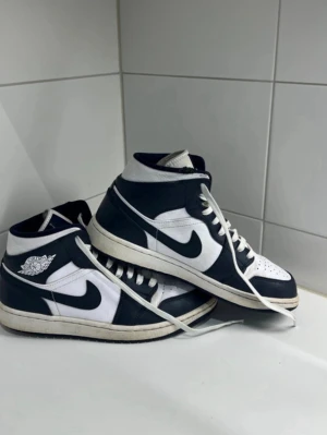 Nike Air Jordan 1 blåvita sneakers - Säljer ett par klassiska Nike Air Jordan 1 sneakers i börkblå och vitt. Skorna har hög siluett, snörning och den ikoniska swoosh-loggan på sidan. Tillverkade i skinn med perforerad tå och retro vingsymbol på ankeln. Perfekta för dig som gillar streetwear och tidlös stil.