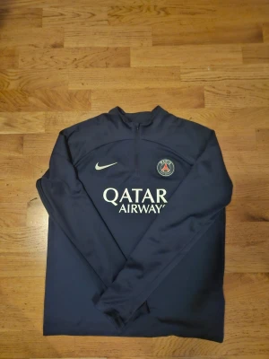 Mörkblå PSG träningströja Nike - Snygg mörkblå långärmad träningströja från Nike med PSG-logga och Qatar Airways-tryck på bröstet. Tröjan har halv dragkedja framtill och är gjord i ett lätt, sportigt material som passar perfekt till träning eller uppvärmning.
