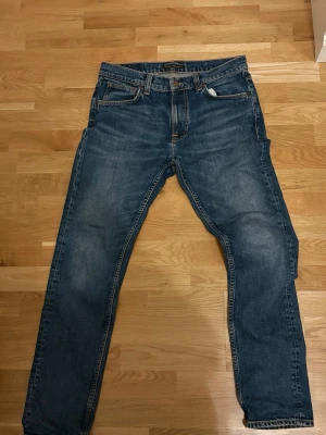 Blå jeans från Nudie Jeans - Säljer ett par klassiska blå jeans från Nudie Jeans med raka ben och fem fickor. Jeansen har kontrastsömmar och Nudie-loggan broderad på bakfickorna. Perfekta till vardags och har en tidlös look.