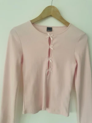 Rosa ribbad topp med rosetter Gina Tricot - Supersöt ljusrosa ribbad topp från Gina Tricot med långa ärmar och tre dekorativa rosetter framtill. Toppen har en tight passform och är perfekt för dig som gillar gulliga detaljer och en feminin stil.