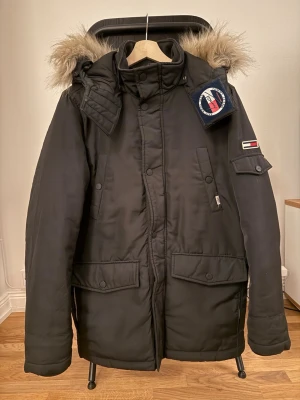 Svart vinterjacka från Tommy Hilfiger - Svart vinterjacka från Tommy Hilfiger med avtagbar huva och fuskpäls. Jackan har stora fickor framtill, patch med logga på bröstet och ärmen samt dragkedja och knappar. Perfekt för kalla dagar med sin varma och rejäla design.
