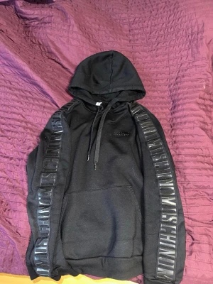 Svart hoodie från Moschino - Snygg svart hoodie från Moschino med huva och dragsko. Stora Moschino-loggor i blankt tryck längs båda ärmarna och mindre logga på bröstet. Klassisk känguruficka framtill. Perfekt för dig som gillar streetwear och vill sticka ut. Storlek S men passar även S/M