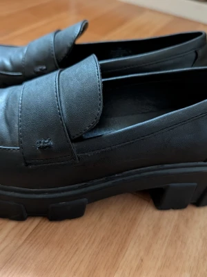 Svarta chunky loafers Claudia Ghizzani - Stilrena svarta loafers från Claudia Ghizzani med chunky sula och klassisk pennyloafers-design. Skorna är tillverkade i syntetmaterial och har en rund tå samt markerad sula för en modern look. 