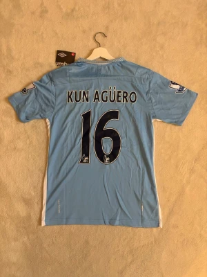 Aguero matchtröja - Snygg ljusblå Manchester City matchtröja från Umbro med 'KUN AGÜERO' och nummer 16 på ryggen. Tröjan har vita detaljer på sidorna, klubbmärke och sponsortryck på bröstet samt Premier League-loggor på ärmarna. 
