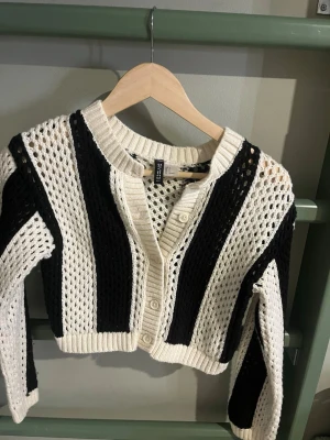 Randig virkad kofta från H&M Divided - Säljer en svartvit virkad kofta från H&M Divided. Koftan har breda ränder, knappstängning framtill och är croppad i modellen. Materialet är grovt stickat med hålmönster och ribbade kanter vid ärmslut och nederkant. Perfekt för en trendig lager-på-lager-look.