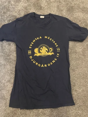 Djurgårdens IF mästartröja 2019 - Mörkblå t-shirt med gult tryck från Djurgårdens IF. Framsidan har texten 'Svenska Mästare 2019' och klubbens emblem i mitten. Rund halsringning och klassisk passform. Perfekt för dig som vill visa ditt stöd för laget.