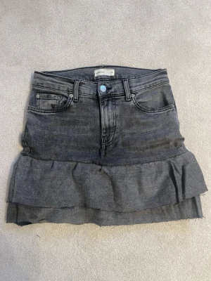 Grå volangkjol i denim från Gina Tricot - Säljer en grå jeanskjol från Gina Tricot med volangdetalj nedtill. Kjolen har klassisk femficksdesign, knapp och dragkedja framtill samt rå kant längst ner. Perfekt för dig som gillar en edgy och trendig stil.