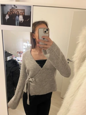 Grå omlottstickad tröja med knyt - Mysig grå stickad omlott-tröja med knyt i sidan och långa ärmar. Tröjan har en avslappnad passform och är perfekt för lager-på-lager. Materialet ser ut att vara en mjuk ull- eller akrylblandning.