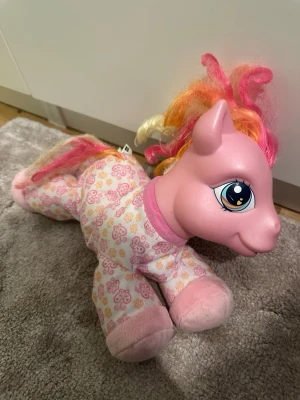 My Little Pony - Söt My Little Pony mjukdjur i rosa med stora blå ögon och färgglad man i rosa, orange och gul. Kroppen har en blommig pyjamas i rosa och orange toner. Perfekt för samlare eller som mysig leksak. Huvudet och frambenen är i hårdplast, de ande sär mjukt. 