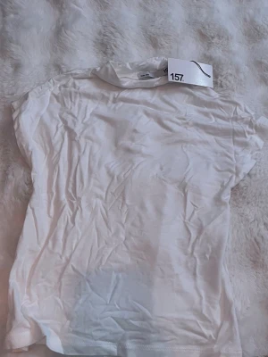 Vit basic t-shirt från 157 - En klassisk vit t-shirt från 157 med rund hals och korta ärmar. Tillverkad i mjuk bomull, perfekt för en enkel och clean look. Passar till allt och är ett måste i garderoben. Den är lite skrynklig och liten storlek men är super dragbar 