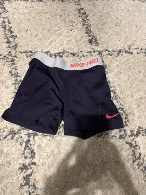 Nike Pro  träningstights-shorts - Nike Pro shorts i mörk lila med bred grå midjeresår och rosa logga. Perfekta för träning, med tight passform och stretchigt material som ger bra rörelsefrihet. Snygg och sportig look med Nike Pro-tryck i rosa på linningen. OBS BARN STORLEK MEN PASSAR MIG SOM ÄR 170lång