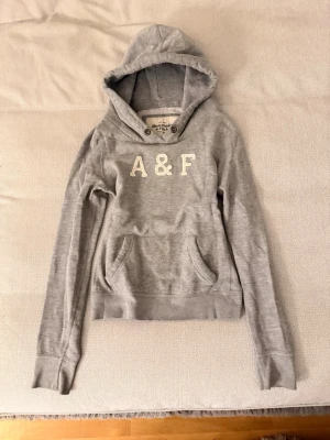 Grå hoodie från Abercrombie & Fitch - Snygg grå hoodie från Abercrombie & Fitch med broderad A&F-logga på bröstet. Klassisk känguruficka framtill, huva med dragsko och långa ärmar. Perfekt för en avslappnad och trendig stil. Mjuk bomullsblandning för extra komfort.