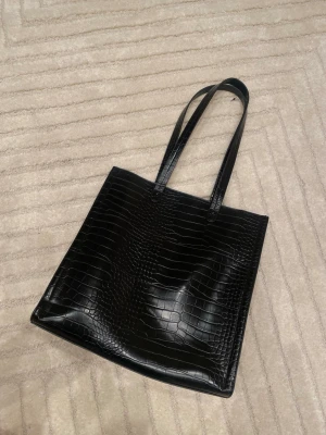 Svart tote-väska från Kappahl – croco-mönster - Elegant och rymlig svart tote-väska från Kappahl i stilrent croco-mönster. Perfekt både till vardag, jobb eller studier. Väskan har långa handtag så den är bekväm att bära över axeln.  Invändigt finns gott om plats samt en innerficka med dragkedja som gör det enkelt att hålla ordning på mindre saker. Väskan stängs med dragkedja upptill.  ✔ Tidlös design ✔ Rymlig och praktisk ✔ Passar laptop, böcker och vardagsnödvändigheter ✔ Bra skick, sparsamt använd  Färg: Svart Märke: Kappahl