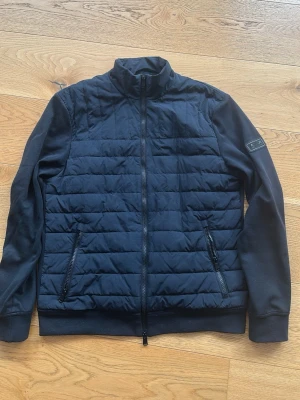 Barbour cardigan med dragkedja - Svart barbour cardigan med quiltad front och släta ärmar. Jackan har hög krage, två fickor med dragkedja och ribbade muddar. Materialmix av polyester och bomull för en skön känsla. Perfekt för lager-på-lager och enkel att matcha. Den är i bra skick, inga skavanker eller liknande. Nypris ca 2500kr. Hör av er vid funderingar!