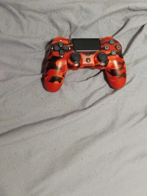 Röd kamouflage PS4 handkontroll - Säljer en snygg röd kamouflage PlayStation 4 handkontroll från Sony. Kontrollern har svart och rött camomönster, klassisk DualShock 4-design med touchpad, två styrspakar och alla vanliga knappar. Perfekt för gamingkvällar!