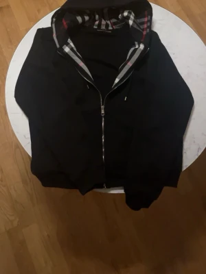 Svart zip-hoodie från Burberry XL - Svart hoodie från Burberry med dragkedja och klassiskt rutigt foder i huvan. Långa ärmar, fickor framtill och justerbar huva med snören. Perfekt för dig som gillar exklusiv streetwear och vill ha något som sticker ut.
