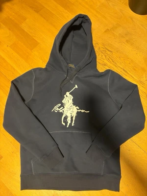 Mörkblå hoodie från Polo Ralph Lauren - Marinblå hoodie från Polo Ralph Lauren med stor vit logga på bröstet. Klassisk modell med huva och dragsko, samt känguruficka framtill. Tillverkad i mjukt bomullsmaterial och har ribbade muddar vid ärmslut och nederkant.