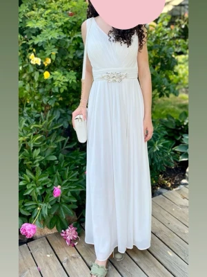 Vit långklänning med dekorerat midjeband - Elegant vit långklänning med v-ringad omlottdesign och dekorerat midjeband med pärlor och broderi. Klänningen är ärmlös och har ett flytande, luftigt fall. Perfekt för speciella tillfällen och studentfirande, bal mm. Skulle uppskatta att den är mellan storlekarna xs/s. Klänningen är köpt i en butik i marbella Spanien
