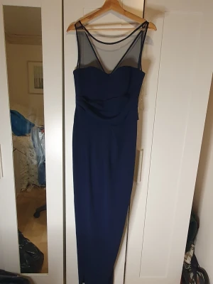 Marinblå långklänning med meshdetalj - Snygg marinblå långklänning med transparent mesh upptill och transparent tyg ovantill.. Klänningen har en figurnära passform och en elegant, enkel design utan mönster med hög slits. Perfekt för dig som vill ha en stilren look med en modern twist.