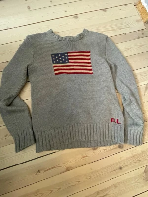 Grå stickad tröja Polo Ralph Lauren - Grå stickad tröja från Polo Ralph Lauren med amerikansk flagga på bröstet och RL-broderi i rött vid nederkanten. Tröjan har rund halsringning och ribbstickade muddar vid ärmslut och nederkant. Perfekt för dig som gillar klassisk college-stil.         Storlek L(14-16)
