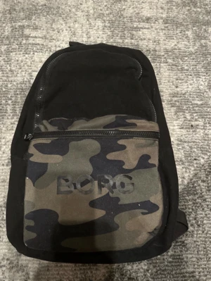Svart ryggsäck från BJÖRN BORG med camo - Snygg svart ryggsäck från BORG med ett stort fack och en framficka i camouflage-mönster. Justerbara axelband och dragkedja framtill. Perfekt för skolan eller häng med kompisar. Logga på framfickan.