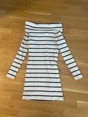 Randig offshoulder klänning - Säljer en vit ribbad kortklänning med svarta horisontella ränder. Klänningen har offshoulder-design och långa ärmar, vilket ger en trendig och avslappnad look. Perfekt för dig som gillar stilrena och moderna plagg.