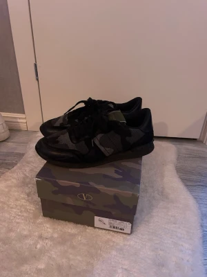 Valentino Rockrunner camo sneakers - Snygga Valentino Rockrunner sneakers i svart och grått camouflagemönster. Skorna har snörning, rund tå och platt sula. Tillverkade i en mix av skinn och textil för en exklusiv känsla. Perfekta för dig som vill ha en unik och trendig look. Skriv om ni undrar något