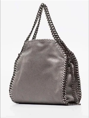 Grå Stella McCartney Falabella väska tiny - Snygg grå handväska från Stella McCartney i modellen Falabella. Väskan har kedjedetaljer längs kanterna och dubbla kedjehandtag. Tillverkad i veganskt läder med en mjuk, lyxig känsla och cool design. Perfekt för dig som gillar statement-accessoarer. Väskan har defekt men inger jag tycker märkt när man bär den.