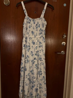 Vit långklänning med blå blommor - Långklänning i vit bomull med blått blommönster och smockad överdel. Klänningen har breda axelband och faller i flera lager för en luftig känsla. Perfekt för varma dagar och ger en romantisk vibe.