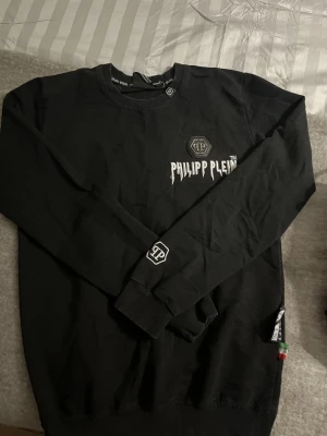 Svart sweatshirt från Philipp Plein - Snygg svart sweatshirt från Philipp Plein med logga och text på bröstet samt en liten logga på ärmen. Tröjan har rund halsringning, ribbade muddar och en diskret etikett i sidan. Perfekt för dig som gillar streetwear och exklusiva detaljer.