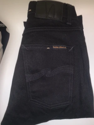 Svarta jeans från Nudie Jeans - Snygga svarta jeans från Nudie Jeans med klassisk femficksdesign och diskret logga på bakfickan. Jeansen har raka ben och är tillverkade i slitstarkt bomullstyg med en mörk tvätt. Perfekta för en stilren look.