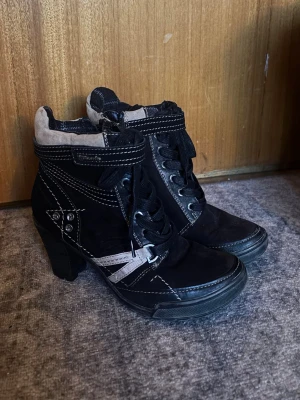 Svarta klackar från Tamaris - Snygga svarta klackar från Tamaris med vita kontrastsömmar, snörning och dekorativa nitar. Skorna har hög klack, rund tå och detaljer i mocka och syntet. Bra skick. 