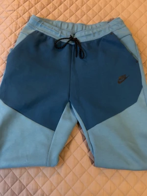 Nike blå mjukisbyxor med snörning (Nike tech) - Snygga blå mjukisbyxor från Nike med två olika blå nyanser och svart Nike-logga på benet. Byxorna har snörning i midjan och dragkedjeförsedd ficka bak. Perfekta för chill eller träning.