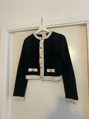 Svart cropped kofta med gulddetaljer - Stilren svart cropped kofta med vita ribbade kanter och guldfärgade knappar framtill. Koftan har rund halsringning, långa ärmar och två dekorativa fickor framtill. Perfekt för att lyfta din outfit med en chic touch.