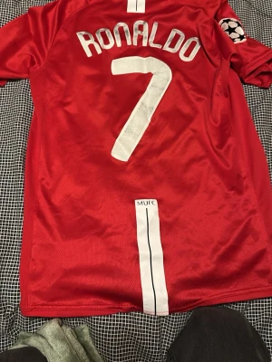 Nästan helt oanvänd Manchester United Ronaldo tröja - Röd Manchester United matchtröja med AIG-logga på bröstet och Ronaldo 7 på ryggen. Tröjan har klubbmärke, Champions League-märke på ärmen och är tillverkad i lätt, ventilerande polyester. Perfekt för fotbollsfans som vill sticka ut på planen eller läktaren.
