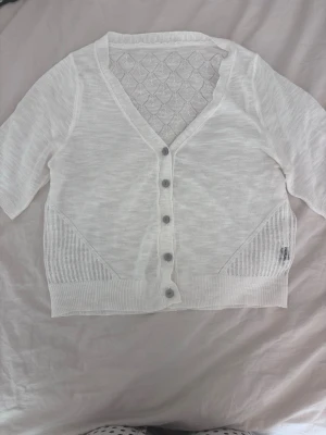 Vit kortärmad kofta med knappar - Snygg vit kofta med korta ärmar och knäppning framtill. Koftan har ett diskret mönster upptill på ryggen och sidorna samt ribbade muddar. Perfekt att slänga över en topp för en fräsch look.