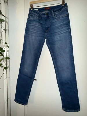 Blå jeans modell Clarke.storlek:Längd 32,Bredd 29 - Klassiska blå jeans från Jack & jones med  raka ben och normal passform. Jeansen har fem fickor, knappgylf och snygga slitningar för en avslappnad look. Perfekta till vardags och lätta att matcha med olika stilar.