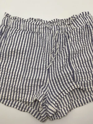 Randiga shorts från Kappahl - Säljer ett par blåvit randiga shorts från Kappahl med bekväm resårmidja och snörning framtill. Shortsen har en avslappnad passform och är tillverkade i ett lätt och luftigt bomullstyg, perfekta för varma dagar. Andvända 1-3ggr