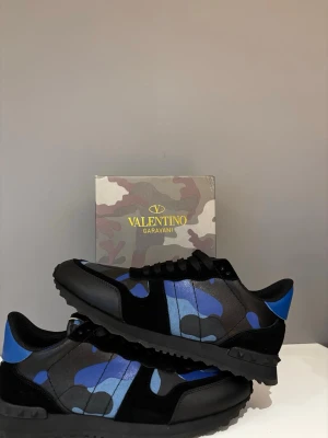 Valentino Garavani blå camo sneakers - Snygga sneakers från Valentino Garavani med blå och svarta camouflage-detaljer. Skorna har svarta snören, rund tå och platt sula. Materialmix av skinn och textil ger en exklusiv känsla. Perfekta för dig som vill sticka ut med stilrena och trendiga sneakers.