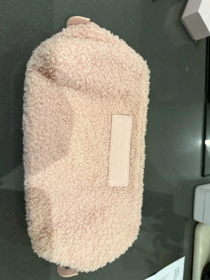 Ljus rosa  necessär Jasmine x CAIA - Söt och fluffig necessär från Jasmine x CAIA. Tillverkad i mjukt teddy-liknande material med en enkel rektangulär form och diskret logga på framsidan. Perfekt för att förvara smink eller småprylar i väskan.